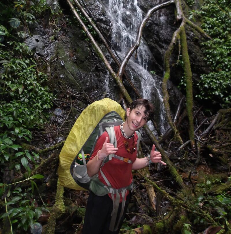 A person with a backpack stands in front of a waterfall. The person is wearing a red shirt and dark pants. They are smiling and making a hand gesture. The backpack is yellow and green. The waterfall is surrounded by lush green vegetation and moss-covered branches. The scene suggests a hike or outdoor adventure in a natural setting.